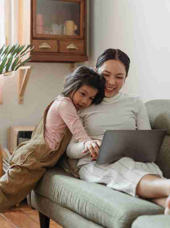 A heartwarming scene of a young child embracing a smiling woman from behind as they both look at a laptop screen together. The woman, likely the mother, is guiding the child's hand on the trackpad, engaging in a shared activity. They are comfortably seated on a couch, surrounded by a homely and well-lit interior that suggests a relaxed and nurturing environment.