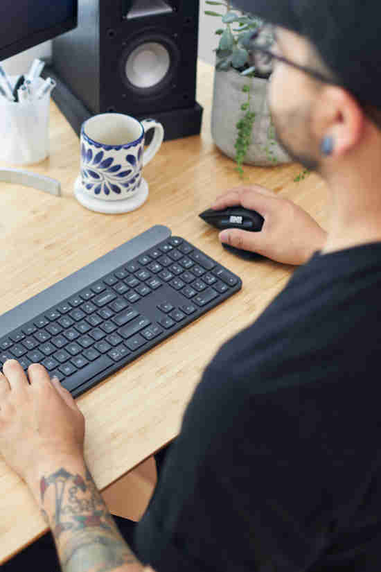 An over-the-shoulder view of a person with a black cap and a black T-shirt working at a computer desk. They are using a black keyboard and mouse on a wooden tabletop. To the left, there's a decorative mug and a pencil holder next to a speaker, with a potted plant adding a touch of greenery to the workspace. The focus on the hands and the computer peripherals suggests a calm and organized work environment.