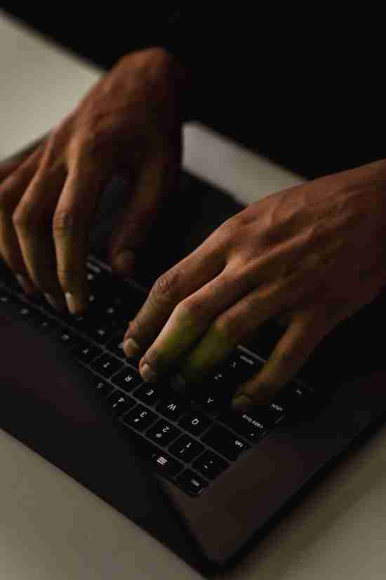 A close-up of two hands typing on a black laptop keyboard, capturing the dynamic motion of fingers pressing keys. The image is set against a high-contrast background, with the dark tones of the laptop and hands starkly outlined by the subtle lighting, emphasizing the action and detail of the typing.