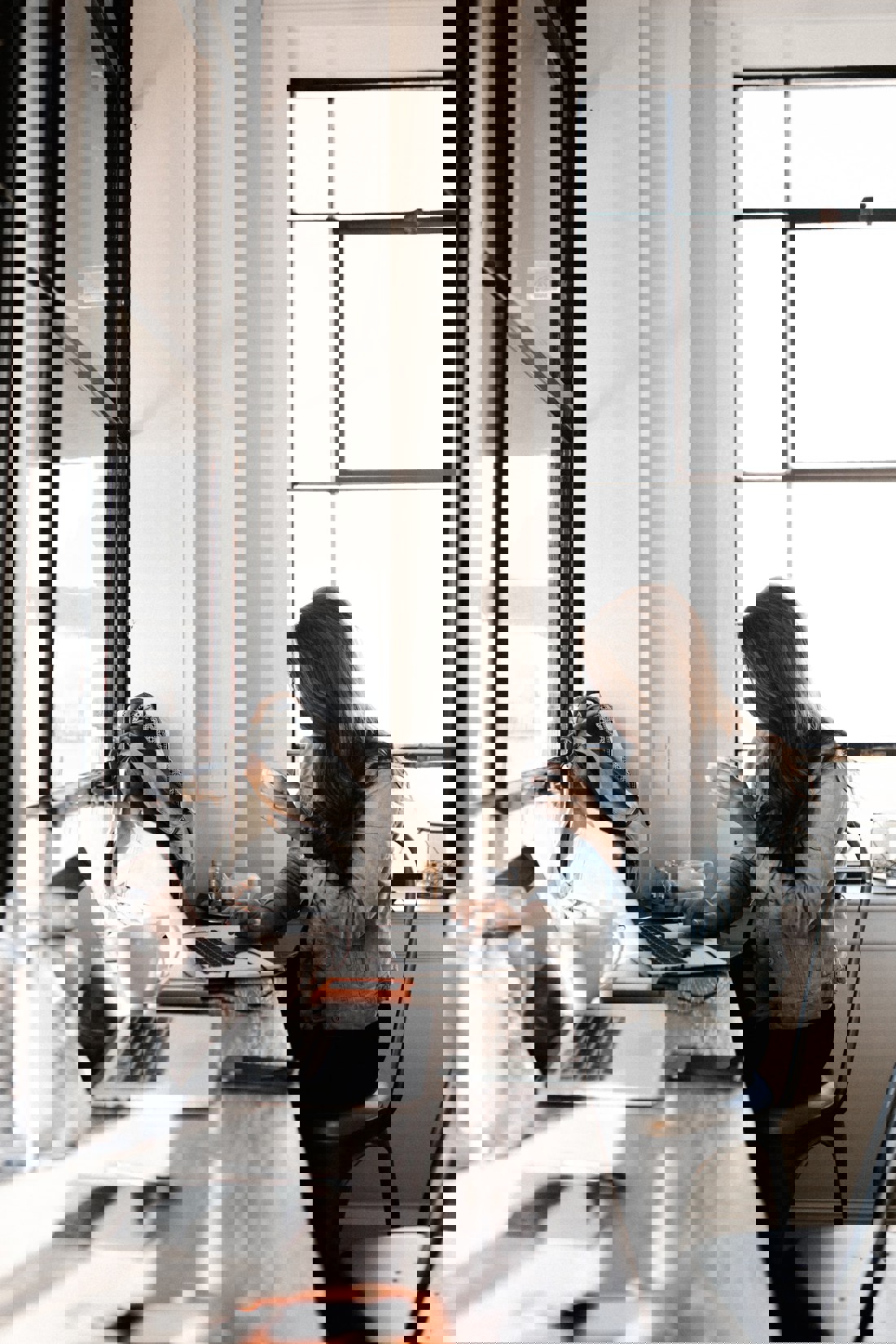 A woman sits at a bar-height table near large windows that fill the room with natural light. She has long hair, wears a denim jacket, and is holding a cup, seemingly taking a break from working on her laptop. Personal items like a backpack and a notebook are scattered on the table, with the urban outline of buildings visible through the window, suggesting a city café or a co-working space.