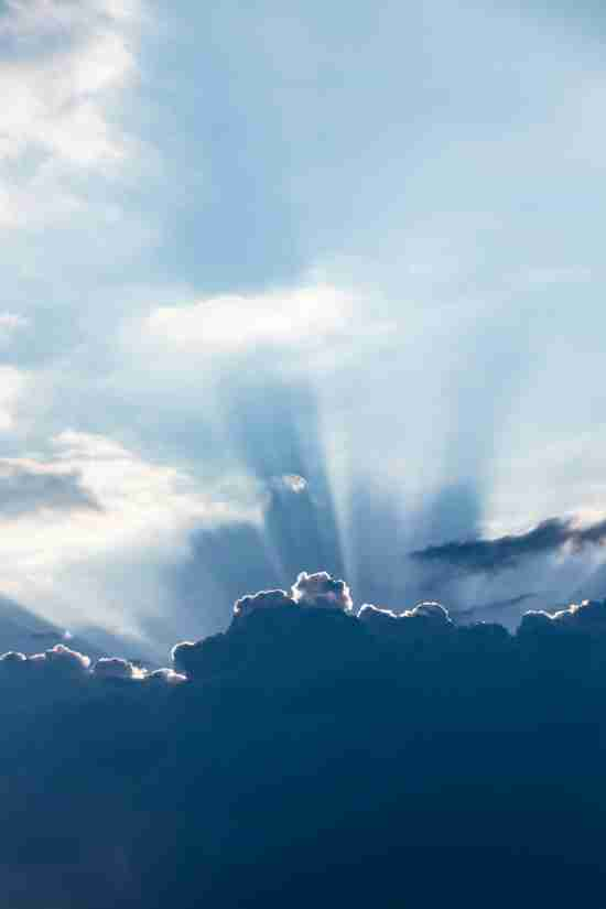 A dramatic sky with sunlight streaming through an opening in the dark, billowing clouds. The rays of light fan outwards, creating a natural spotlight effect against the contrasting shadows of the clouds, evoking a sense of hope and inspiration.