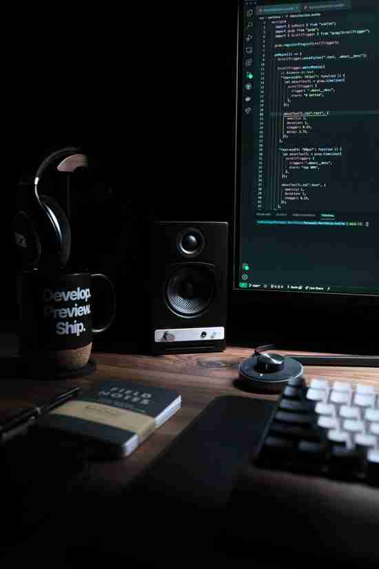 A programmer's workspace illuminated by the soft glow of a monitor displaying code. In the foreground, a mechanical keyboard and a sleek, modern speaker sit on a dark wood desk. A mug with the words 'Develop, Preview, Ship' reinforces the coding theme. Headphones hang on the mug, suggesting a focus on work. A notebook titled 'FIELD NOTES' lies beside a pen, indicating a blend of analog and digital work tools.