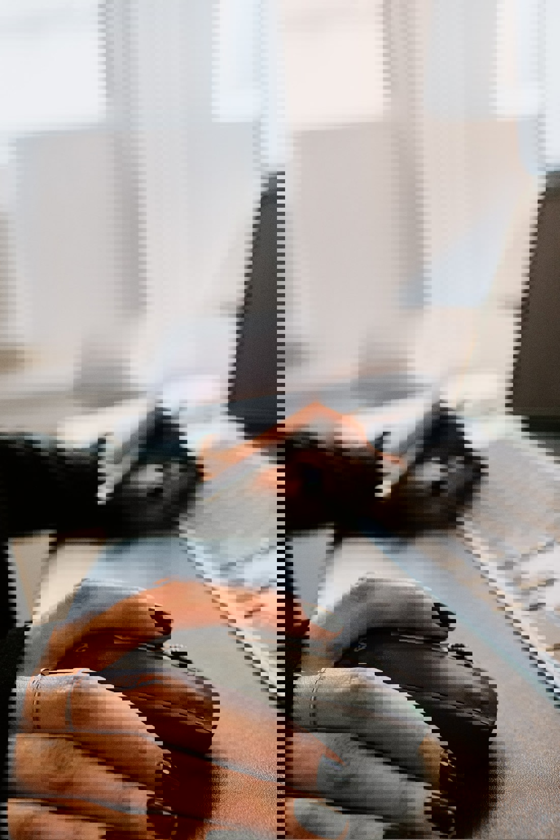 A focused view on a person's hands with black nail polish, one hand resting on a wireless mouse and the other holding a smartphone. The mouse is on a desk beside a laptop with a partial view of the keyboard. The blurred background suggests an indoor setting with soft lighting, providing a glimpse into a typical workspace scenario.