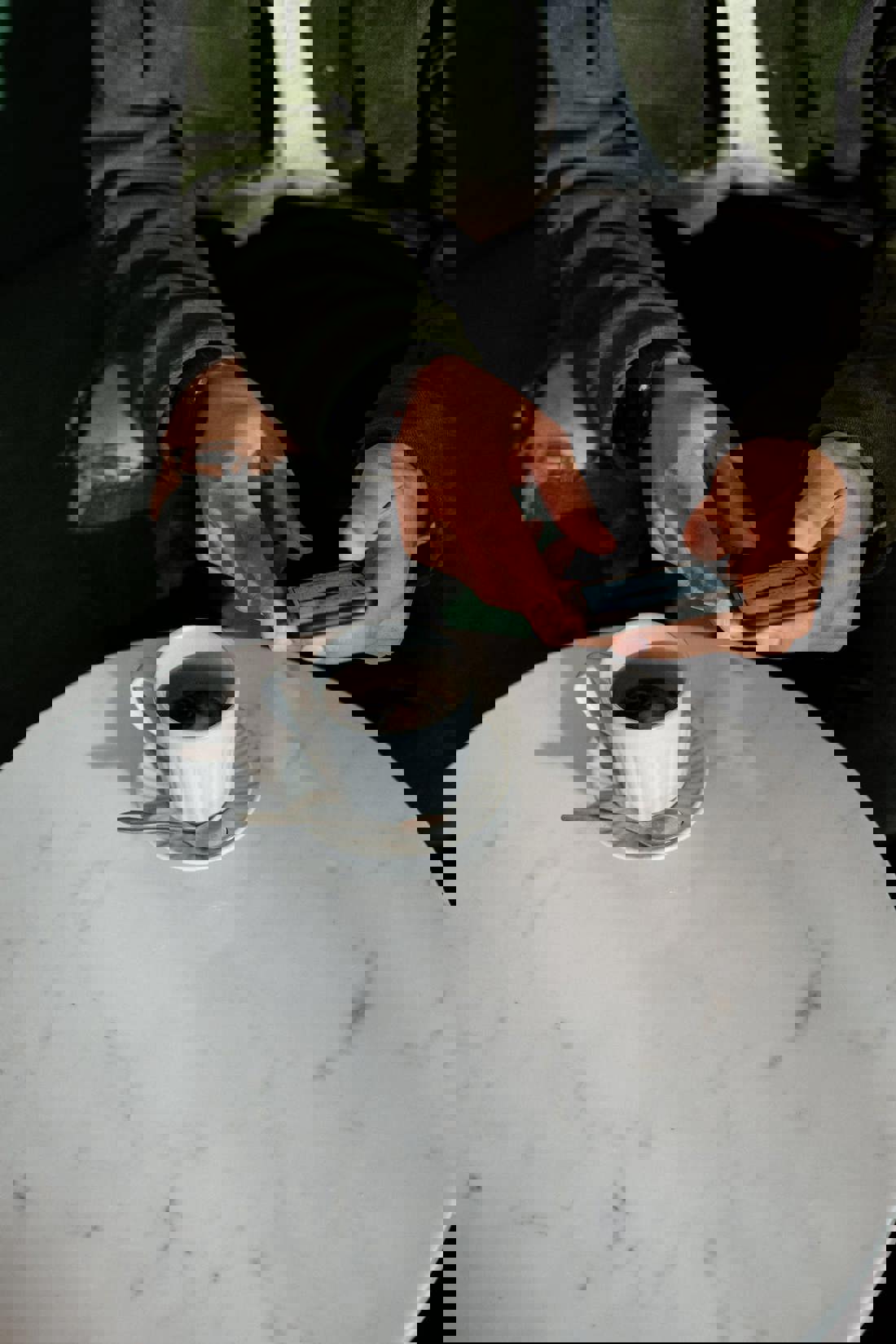 A close-up image showing a person's hands above a white marble table as they use a smartphone. On the table, there's a cup of coffee on a saucer with a spoon beside it. The person is wearing a green jacket with rolled-up sleeves, revealing a tattoo on the forearm, and is seated in a casual, modern setting.