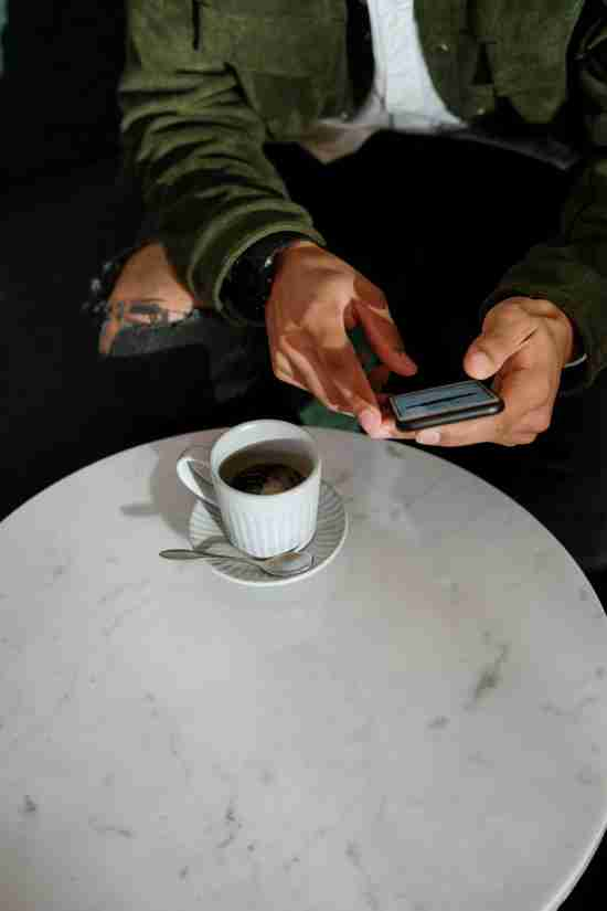 A close-up image showing a person's hands above a white marble table as they use a smartphone. On the table, there's a cup of coffee on a saucer with a spoon beside it. The person is wearing a green jacket with rolled-up sleeves, revealing a tattoo on the forearm, and is seated in a casual, modern setting.
