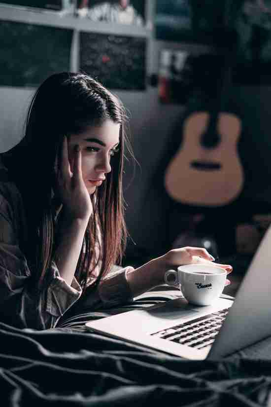 A young woman is seen engaging with a laptop in a dimly lit room, her face illuminated by the screen's glow. She rests her cheek in her hand while holding a white cup with the word 'Cocoon' printed on it, suggesting a moment of relaxation or deep concentration. A guitar hangs on the wall in the background, adding a personal touch to the cosy setting.