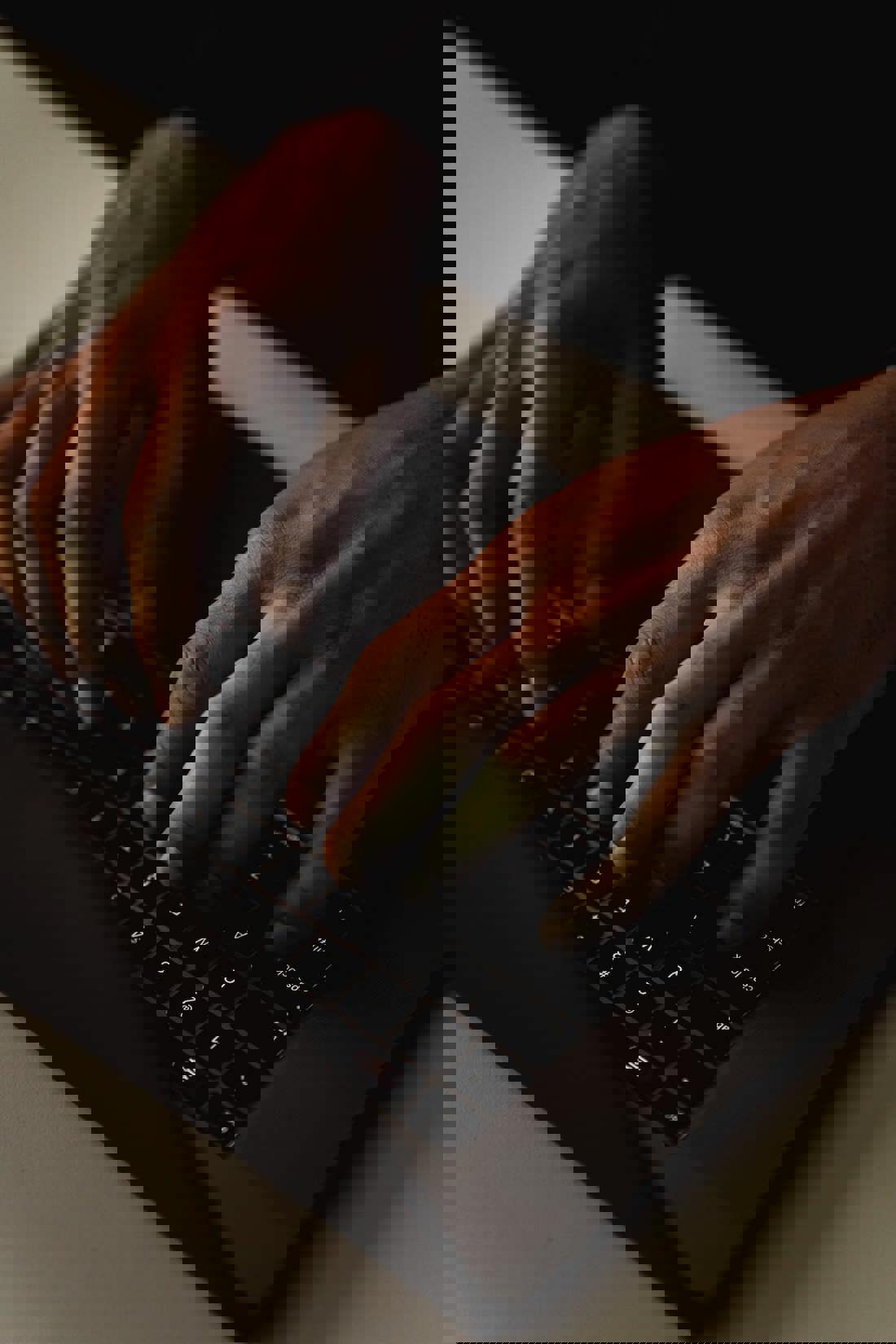 A close-up of two hands typing on a black laptop keyboard, capturing the dynamic motion of fingers pressing keys. The image is set against a high-contrast background, with the dark tones of the laptop and hands starkly outlined by the subtle lighting, emphasizing the action and detail of the typing.