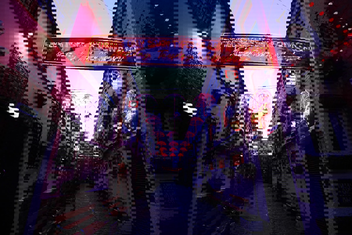 A vibrant alleyway in Belfast during the evening, illuminated by neon signs including one that humorously reads, 'There's only seven types of rain in Belfast: Monday, Tuesday, Wednesday...'. Red umbrella canopies are suspended above the alley, creating a whimsical overhead display. Various signs for Guinness and other establishments line the walls, alongside hanging flower baskets and benches, contributing to the alley's lively ambiance.