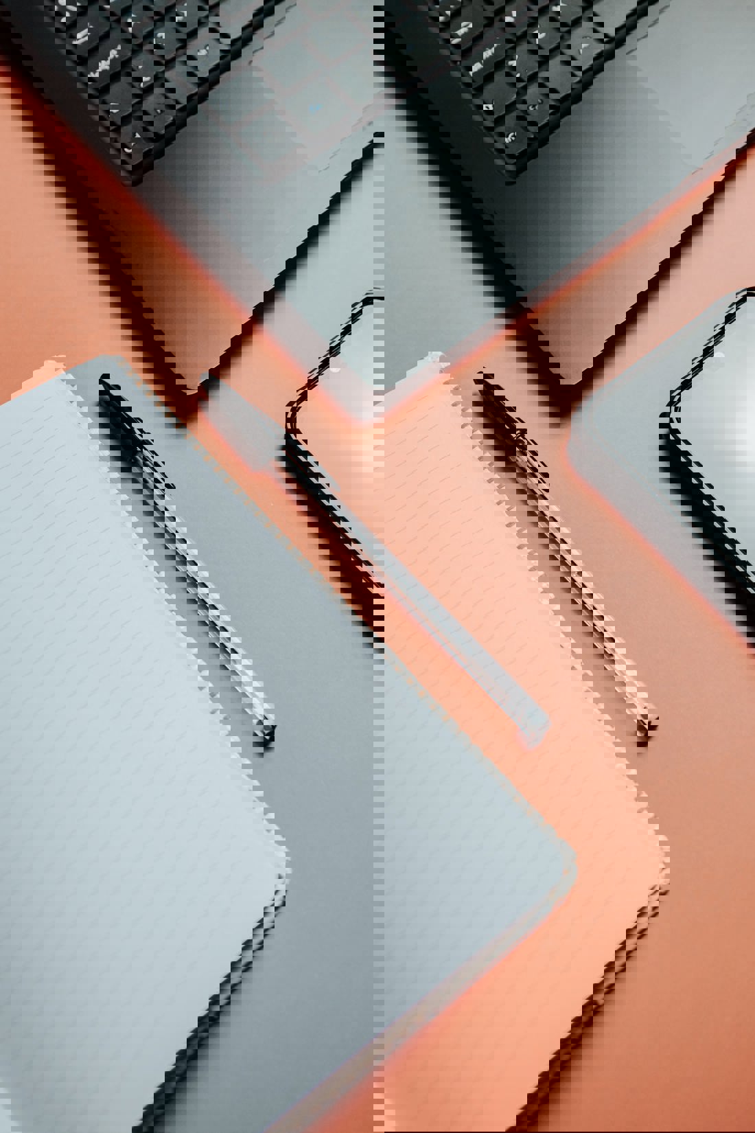 A flat lay photograph of a workspace with a black laptop keyboard in the upper left corner, a lined spiral notebook with a black pen on top in the center, and a smartphone in the lower right corner. All items are placed on a peach-colored surface, suggesting a modern, minimalist work environment.