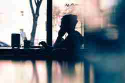 Silhouette of a person sitting at a table in front of a laptop, with a blurred background featuring tree branches and bright daylight coming through a window.