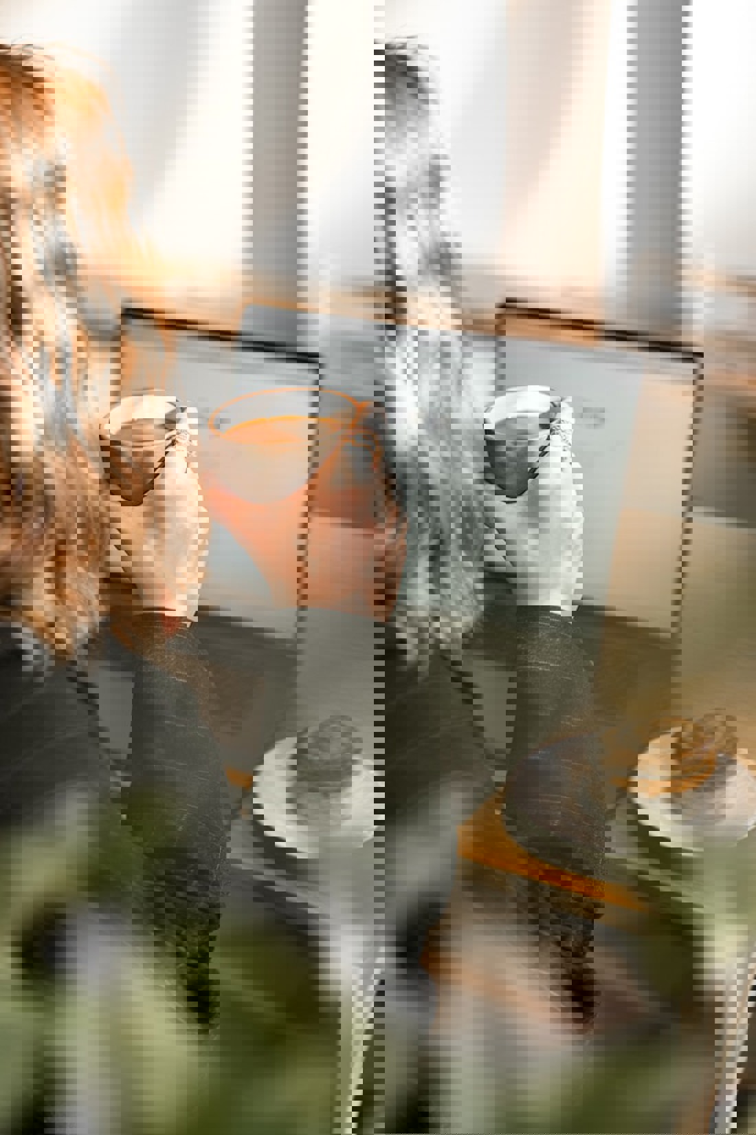 Over-the-shoulder view of a person with wavy blonde hair, sitting in front of a laptop and holding a ceramic cup of coffee. They are wearing a dark jacket with a rolled-up sleeve. On the table beside the laptop is a plate with a cinnamon roll. The environment suggests a comfortable, sunlit space, ideal for work or relaxation, with the presence of greenery in the blurred foreground adding to the cosy atmosphere.